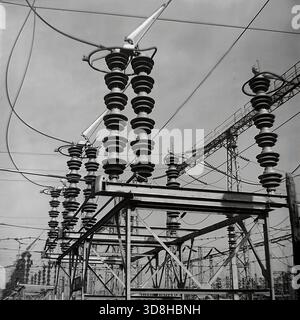 Donetsk region, Ukrainian SSR, Soviet Union. 1970s. Symmetric rows of high-voltage porcelain insulators and disconnectors at the open switchgear of a large electrical substation. The heavy equipment is mounted on metal frames and concrete foundations. In the background, construction cranes and power plant buildings are visible. A worker performs maintenance on an air blast circuit breaker. The geometric patterns of the industrial infrastructure. Life in peaceful Donbas before the war. Stock Photo