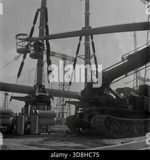 Donetsk region, Ukrainian SSR, Soviet Union. 1970s. A specialized tracked aerial work platform operates at the open switchgear yard of a high-voltage electrical substation. Installers in the basket of the telescopic lift are performing maintenance on massive air blast circuit breakers and porcelain insulators. The heavy crawler chassis allows the machine to maneuver on the rough terrain of the construction site. View through the metal lattice of a transmission tower. Life in peaceful Donbas before the war. Stock Photo