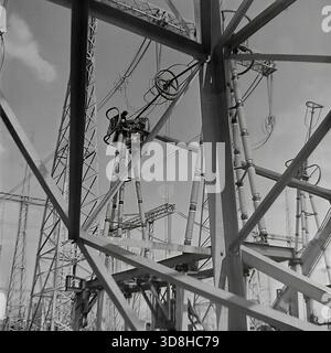 Donetsk region, Ukrainian SSR, Soviet Union. 1970s. A specialized tracked aerial work platform operates at the open switchgear yard of a high-voltage electrical substation. Installers in the basket of the telescopic lift are performing maintenance on massive air blast circuit breakers and porcelain insulators. The heavy crawler chassis allows the machine to maneuver on the rough terrain of the construction site. View through the metal lattice of a transmission tower. Life in peaceful Donbas before the war. Stock Photo