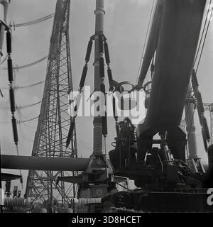 Donetsk region, Ukrainian SSR, Soviet Union. 1970s. A specialized tracked aerial work platform operates at the open switchgear yard of a high-voltage electrical substation. Installers in the basket of the telescopic lift are performing maintenance on massive air blast circuit breakers and porcelain insulators. The heavy crawler chassis allows the machine to maneuver on the rough terrain of the construction site. View through the metal lattice of a transmission tower. Life in peaceful Donbas before the war. Stock Photo