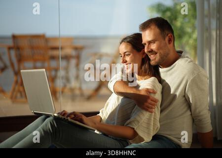 Happy couple in love using laptop sitting on the floor beside a window in a house indoor Stock Photo