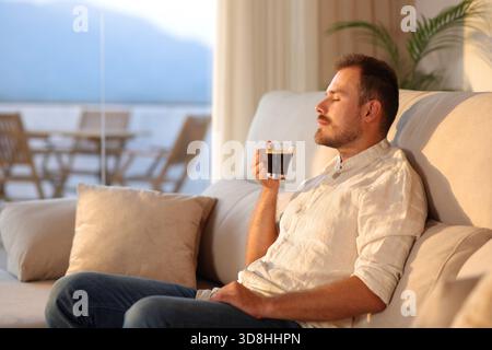 Man smelling coffee sitting on a couch at home at sunset Stock Photo