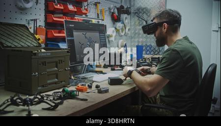 Engineer Using VR Goggles to Virtually Design. Man in Military Style Uniform Utilizing VR Goggles and Computer While Assembling Drone Components. Concept Integration of Virtual Tools in Engineering. Stock Photo