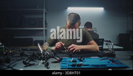 Male Engineer With Tattoos Returns to Desk, Ready to Resume Assembling and Testing FPV Drones. Concept of Focused Nature of High Tech Production and Dedicated Individual Work, Refining drone. Stock Photo