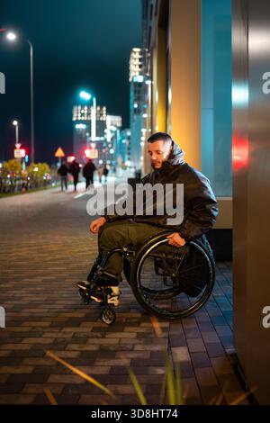 Lonely young disabled man in wheelchair in rgb color light walking in late night city area. Stock Photo