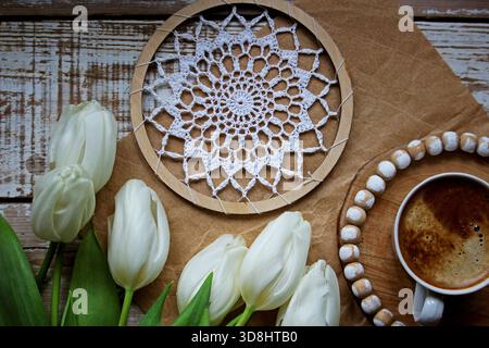 Cozy still life with a cup of coffee, white tulips and handmade crochet lace in a wooden hoop on a rustic wooden background. Stock Photo