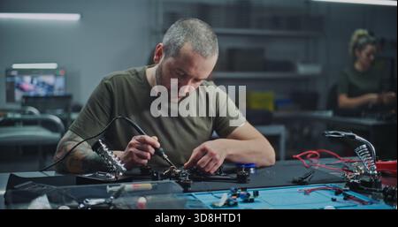 Worker With Tattoo on Arm Soldering Internal Electronics Onto Contemporary Drone Frame in Modern, Industrial Lab. Concept of Intersection of Industrial Design High Tech Assembly, Refining drone. Stock Photo