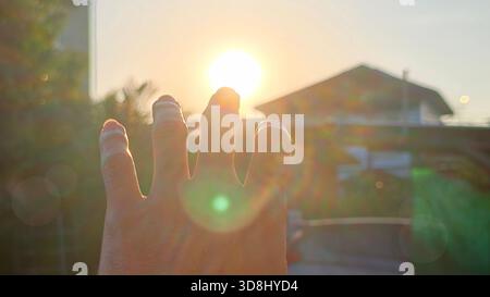 First-person view of a man's outstretched hand wearing, reaching up to cover the bright, setting or rising sun. Features strong backlighting and promi Stock Photo