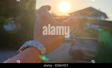 First-person view of a man's outstretched hand wearing, reaching up to cover the bright, setting or rising sun. Features strong backlighting and promi Stock Photo