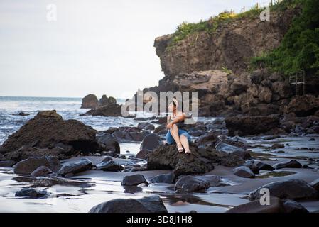 A contemplative woman sits peacefully on a large rock at the beach, gazing out at the ocean Stock Photo