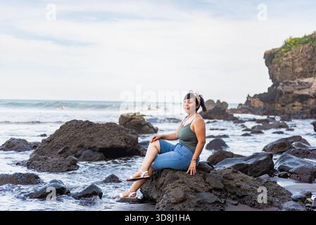 A contemplative woman sits peacefully on a large rock at the beach, gazing out at the ocean Stock Photo