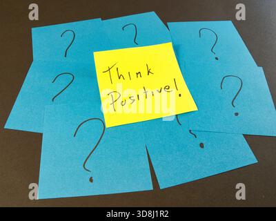 Think Positive! Among Question Marks - Yellow sticky note with 'Think Positive!' surrounded by blue notes with question marks. A concept of optimism, Stock Photo