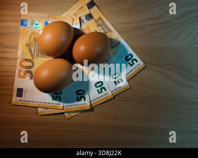 Food Inflation: Eggs and Euro Money - Three brown eggs resting on fanned 50 Euro banknotes on a wooden table. Symbolizes rising food costs, inflation, Stock Photo