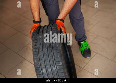 Mechanic with orange gloves moving a tire in the workshop. Car maintenance and tire change for road safety. Stock Photo