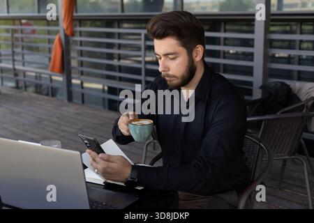 A man is drinking from a mug while using his smartphone. Stock Photo