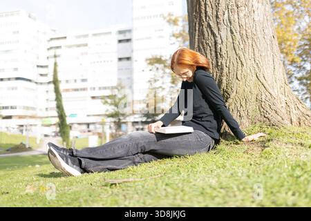 Young woman reading book leaning against tree in park Stock Photo