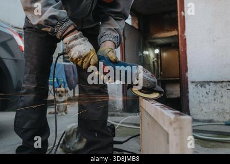 Worker uses angle grinder to cut metal beam outdoors with sparks and dog nearby at a work site Stock Photo