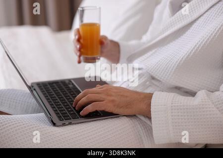 Man in bathrobe with glass of juice working on laptop indoors, closeup Stock Photo