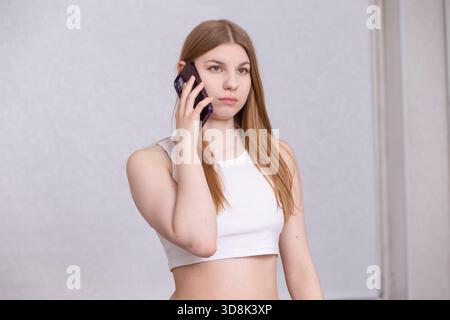 A young woman with long brown hair engages in a phone conversation while standing in a well-lit, neutral-toned indoor area. She appears focused and co Stock Photo