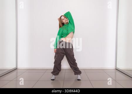 A dancer performs expressive moves in a studio, wearing a green hoodie and loose gray pants. The bright, minimalistic environment enhances her gracefu Stock Photo