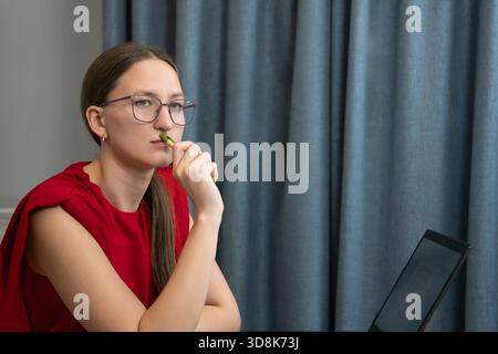 Businesswoman wearing glasses is sitting at desk with laptop and holding pen near her lips, thinking about project and working remotely from home Stock Photo