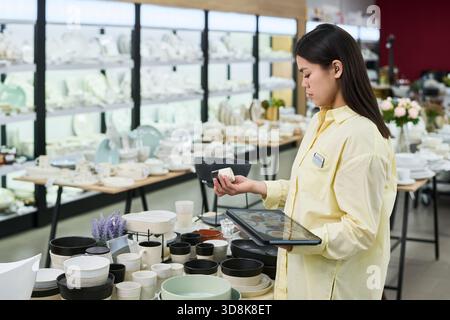 Young woman standing in store holding ceramic cup and digital tablet, checking inventory among shelves filled with tableware and kitchenware products in retail environment Stock Photo