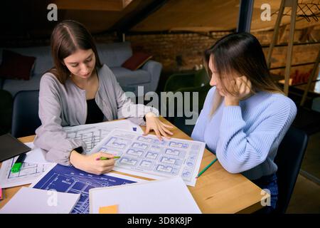 Two young women analyzing architectural drawings during project session Stock Photo