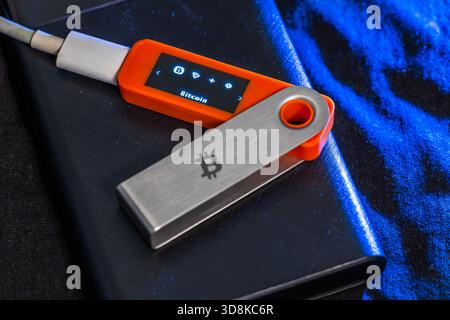 Close-up of cryptocurrency hardware wallet featuring Bitcoin symbol. Silver and orange device connected with cable against dark background, evoking a technological and secure atmosphere. Stock Photo
