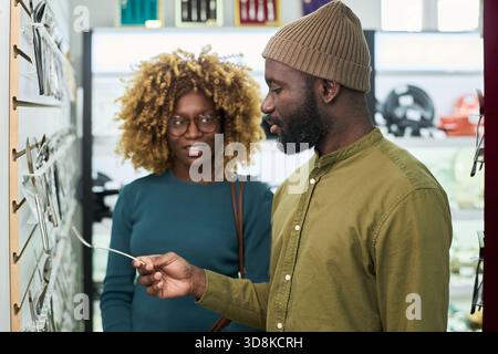 Black man examining eyeglass frames while standing next to Black woman in optical store, both young adults interacting and discussing eyewear selection in retail environment Stock Photo