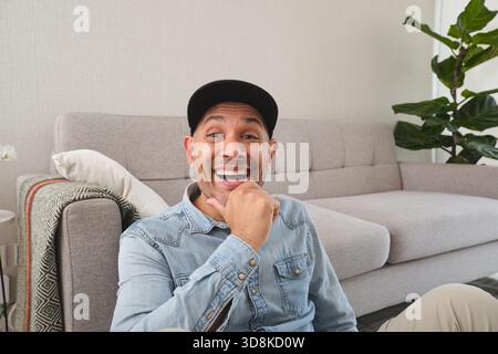 Smiling man wearing baseball cap and denim shirt laughing in living room Stock Photo