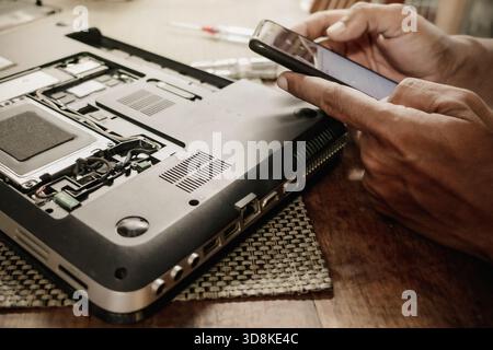 A man trying to repair his broken laptop by searching for guidance from the internet using smartphone. DIY concept. Stock Photo