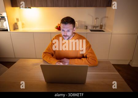 Adult businessman freelancer works at home online on laptop, sitting in kitchen late at night. Stock Photo