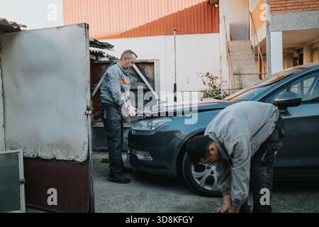 Two technicians repair a car in an outdoor lot, focusing on the hood and tools Stock Photo