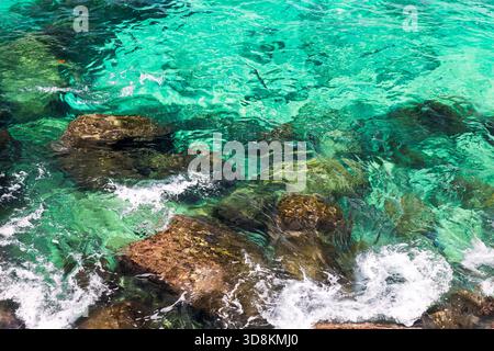 Bright turquoise water flows around rugged rocks, creating a vivid coastal scene. Clear sea reveals underwater textures, while white foam and waves ad Stock Photo