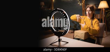 Female blogger unpacking box recording video using smartphone, ring light with tripod making frame with hands and fingers late evening at home office. Stock Photo