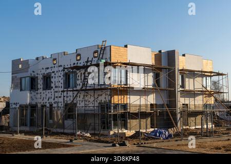 An unfinished two-story residential building with exposed white facade insulation, scaffolding, and wood trim is under construction on a sunny day. Stock Photo
