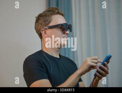 Man wearing modern AR glasses and using a smartphone controller. Futuristic wearable tech, mixed reality device, digital innovation and immersive tech Stock Photo