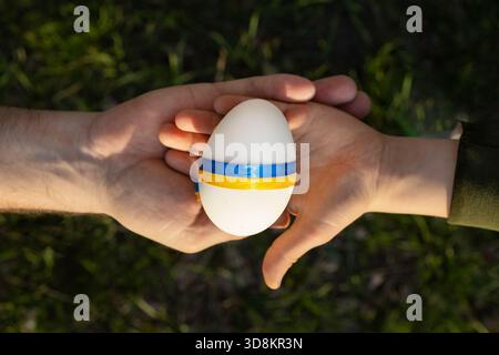 Two hands hold a white Easter egg, tied with yellow and blue ribbons, the colors of the Ukrainian flag. Space for text. Faith in peace, a bright futur Stock Photo