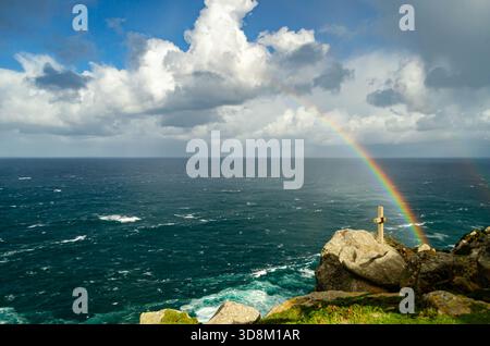 Stunning rainbow arching over a vast ocean with a cross on a rocky cliff. Cloudy blue sky above. Concept of hope faith spirituality memorial and resil Stock Photo
