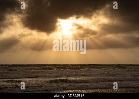 Coastal waves breaking with a sun showing through clouds at sunset casting light rays out from the sky. Stock Photo