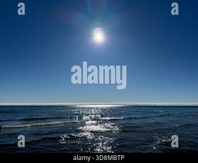 A brilliant sun with visible lens flare glows over calm blue water, creating a serene and reflective mood across gentle waves. Stock Photo
