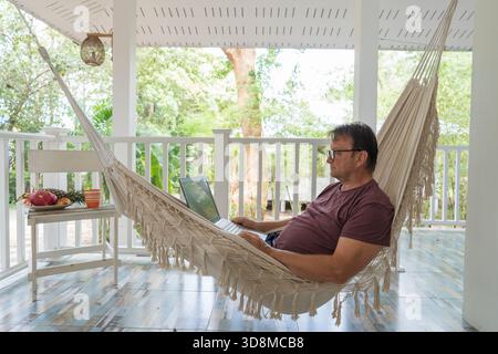 Grown man rests and working a laptop in a hammock on a terrace home near tropical garden in island Koh Phangan, Thailand, close up Stock Photo