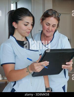 Two female healthcare workers reviewing information together on a black board, smiling and discussing their work in a clinical setting. Stock Photo