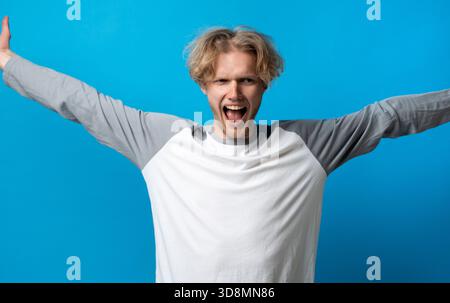 Excited young man raising arms and shouting joyfully on blue background, celebrating success and energy Stock Photo
