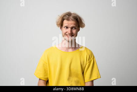 Smiling young man looking sideways with awkward amused expression while posing in neutral studio scene Stock Photo