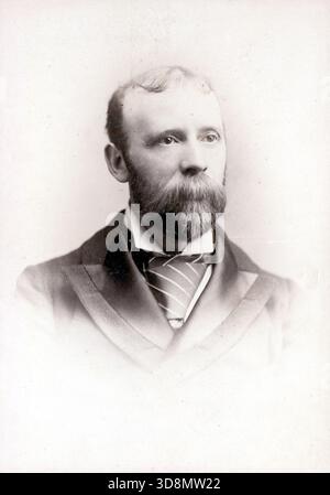 A solemn Victorian gentleman with a full beard and mustache gazes slightly off-camera. His dark coat with wide lapels, crisp white shirt, and striped tie evoke late 19th-century formality. The soft vignette and monochrome palette enhance the introspective mood. Photographed by Embers0on and Sons, distributed through various London outlets. From a late Victorian to early Edwardian original photograph album which featured many albumen cabinet cards and carte de visites, providing a window to the styles and fashions of the time. Stock Photo