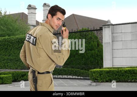 Security guard in uniform using radio near house outdoors. Private property protection Stock Photo