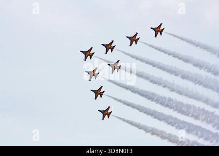 Eight vibrant orange fighter jets fly in tight diagonal formation, trailing long white contrails across a partly cloudy sky at an air show. Stock Photo