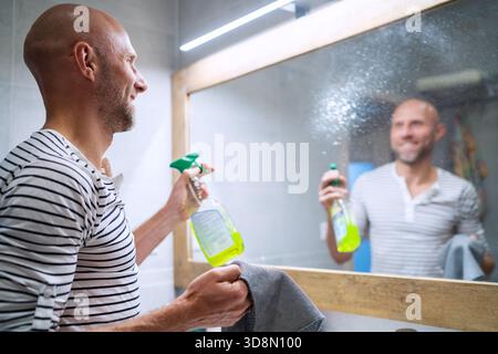 Happy smiling caucasian bald man using detergent sprayer for bathroom mirror cleaning. He spraying it on surface and enjoying cleaning day. Everyday a Stock Photo