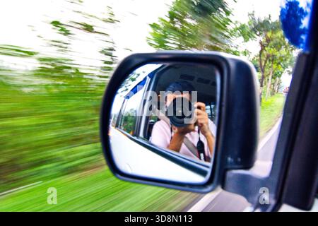 Asian male photographer reflected in speeding van's side mirror photographs motion-blurred lush green rural landscape through open window. Stock Photo